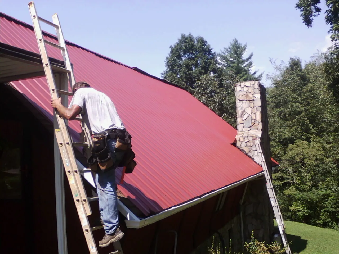Roofer on a ladder beside a red metal roof and stone chimney — Soffit & Fascia Repair by Roofing Brothers in Saint Louisville